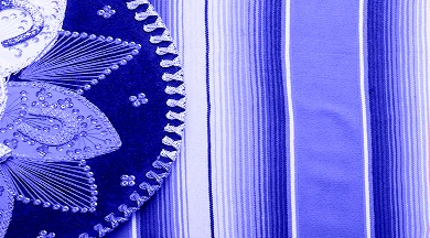 Primer plano con filtro azul de la parte inferior de un sombrero de charro bordado y un sarape mexicano de rayas, representando la exportación de productos locales y la cultura mexicana.
